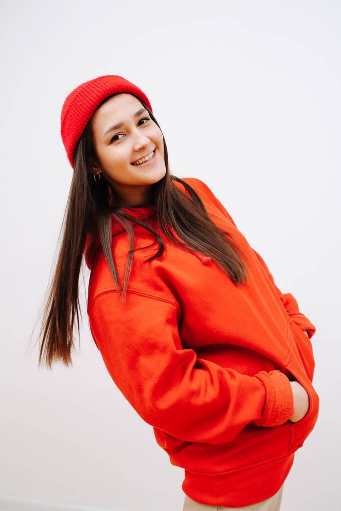 Engaiged smilig woman in vibrant red cap and hoodie tilting back with hands in pockets. She is standing sideways, looking at the camera. Indoors, in front of the white wall. - Φωτογραφία, εικόνα