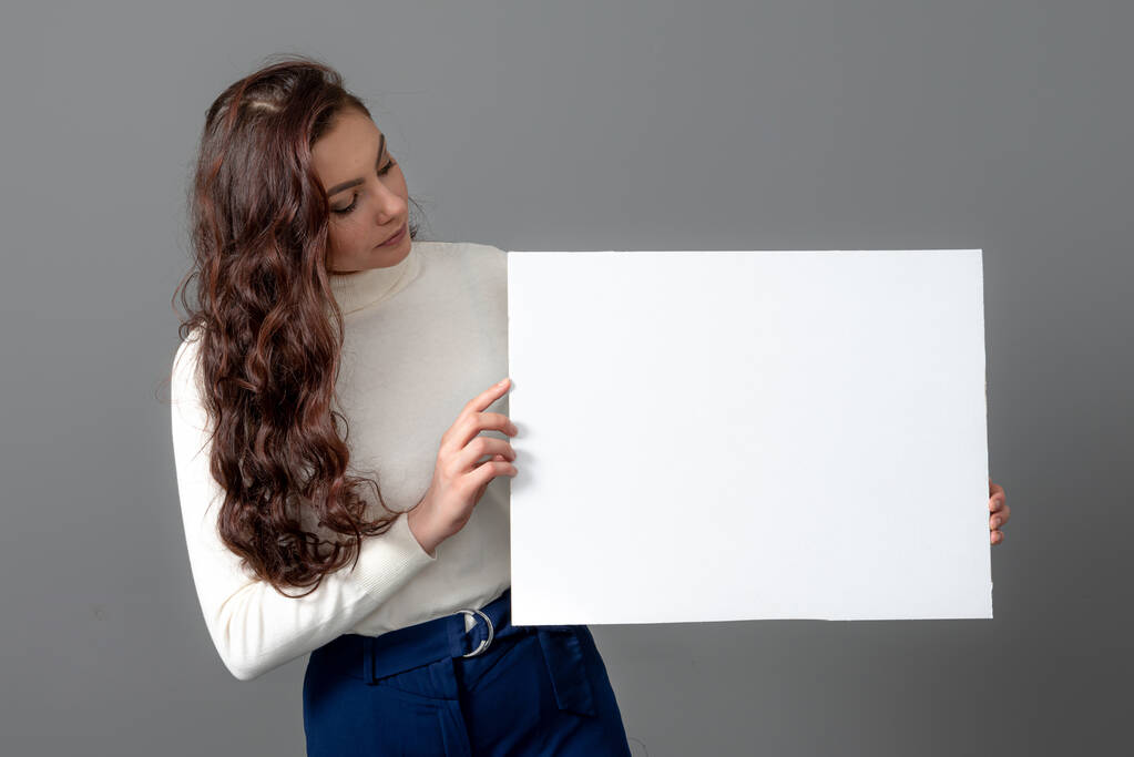 bela mulher de negócios sensual com cabelos longos encaracolados mostra um bilboard em branco, isolado em cinza, espaço de cópia - Foto, Imagem