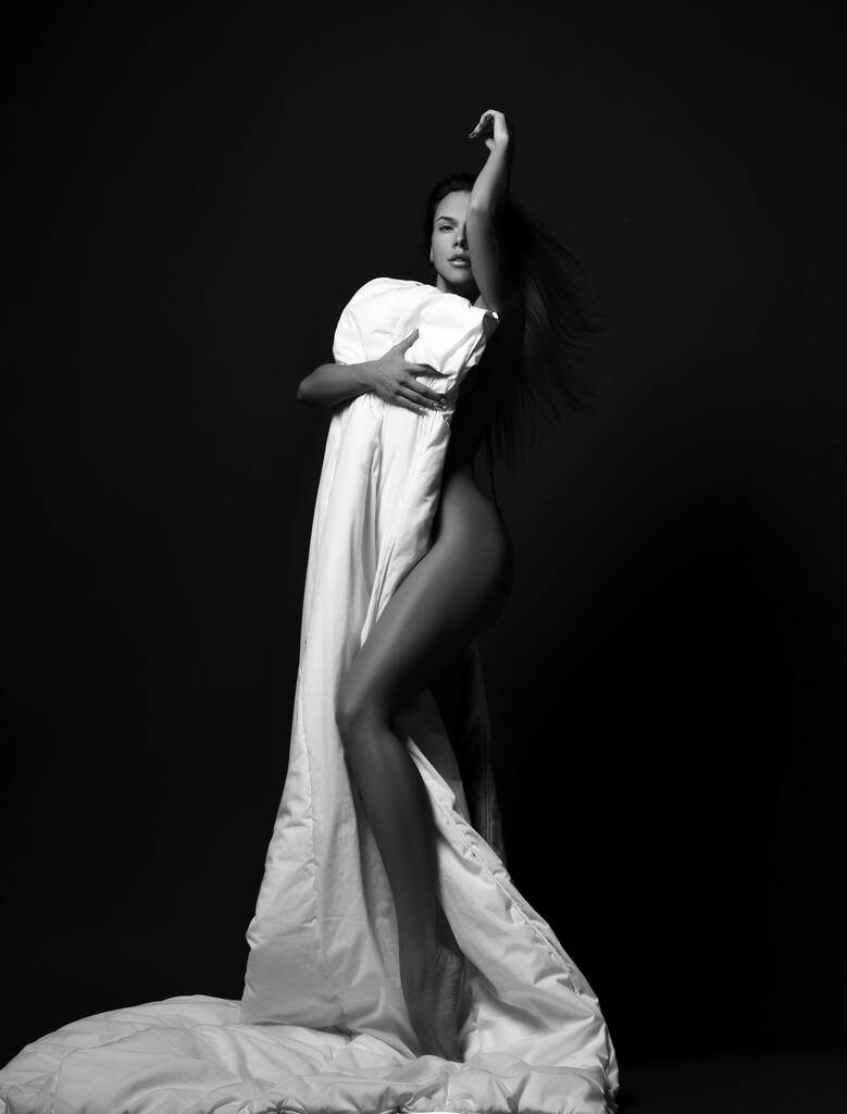 Černobílý. Smyslná sexy nahá žena stojí a zakrývá se velkou měkkou bílou přikrývkou, flirtuje, škádlí - Fotografie, Obrázek