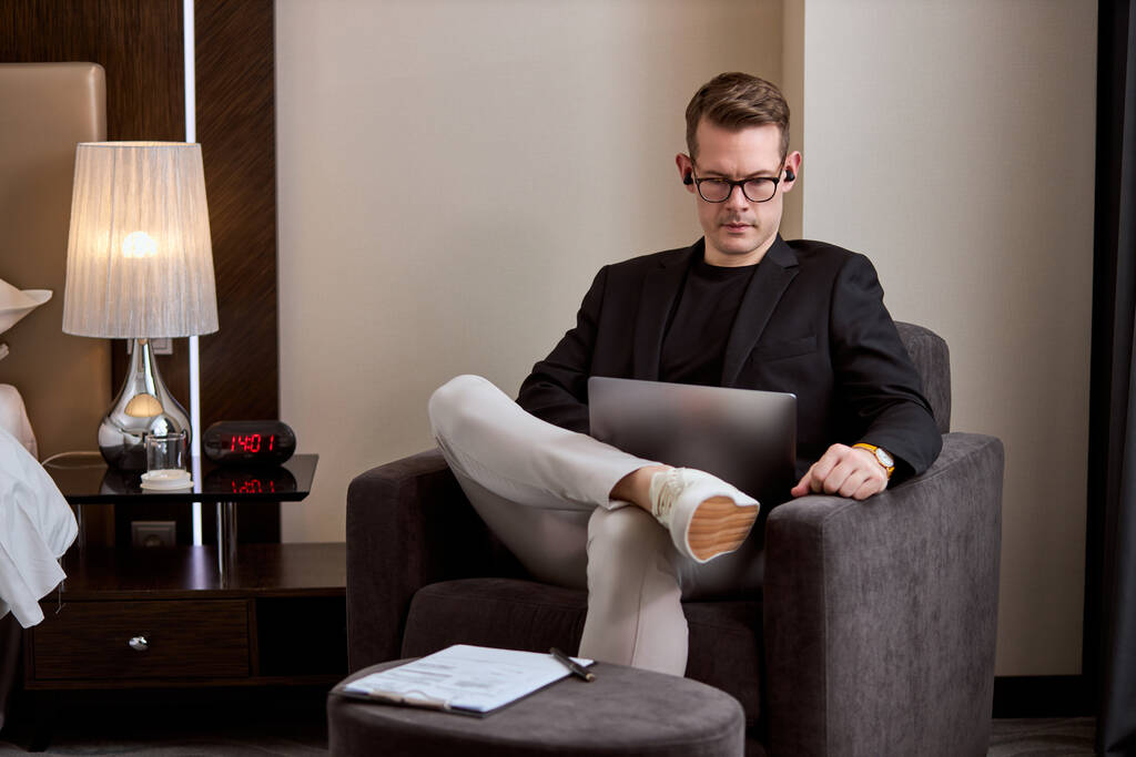 Poważny mężczyzna w eleganckim garniturze siedzi na kanapie pracując na laptopie, nosząc słuchawki bezprzewodowe - Zdjęcie, obraz