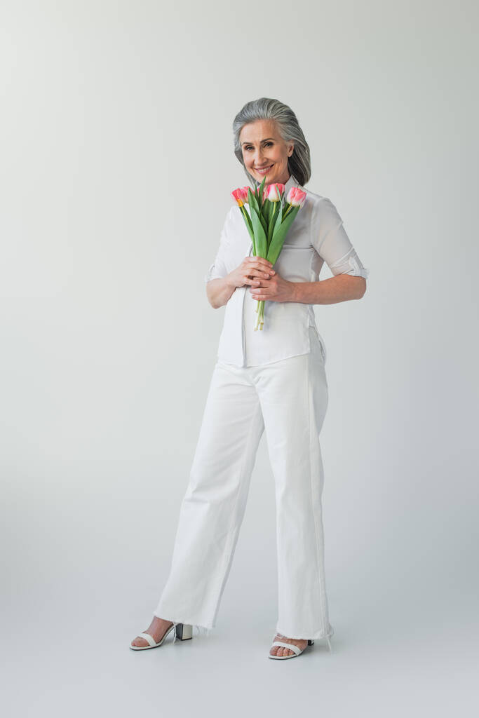 Зрелая женщина улыбается, держа цветы на сером фоне  - Фото, изображение