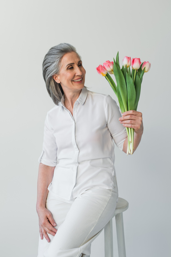 Mujer de pelo gris sonriente mirando ramo de tulipanes aislados en gris  - Foto, imagen