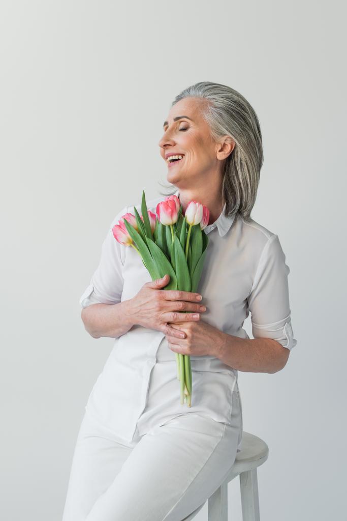 Mature woman with closed eyes smiling while holding tulips isolated on grey  - Photo, Image