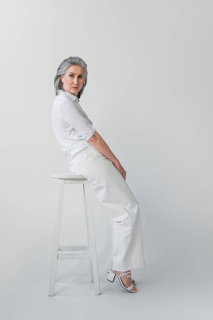 Középkorú nő pózol fehér szék szürke háttér  - Fotó, kép