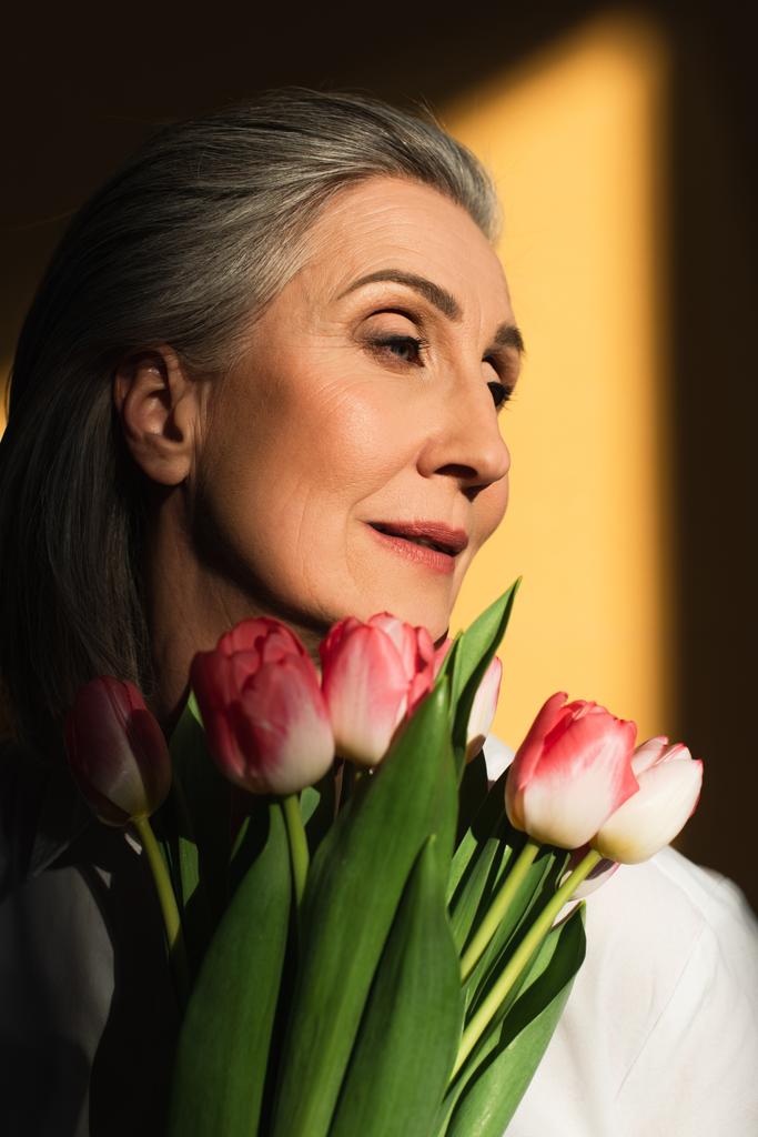 Žena středního věku drží růžové tulipány na žlutém pozadí se stínem  - Fotografie, Obrázek