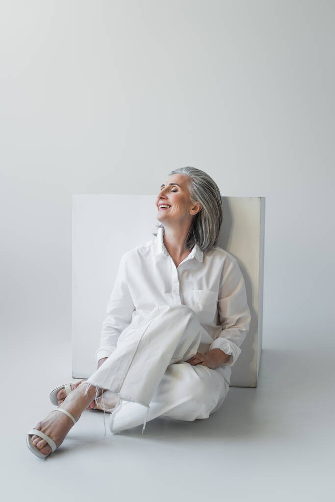 Fröhliche reife Frau mit geschlossenen Augen sitzt neben Würfel auf grauem Hintergrund  - Foto, Bild