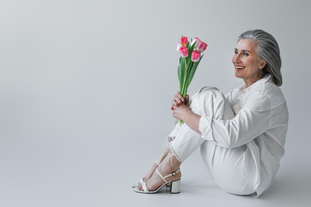 Cheerful mature woman holding bouquet of tulips while sitting on grey background  - Photo, Image