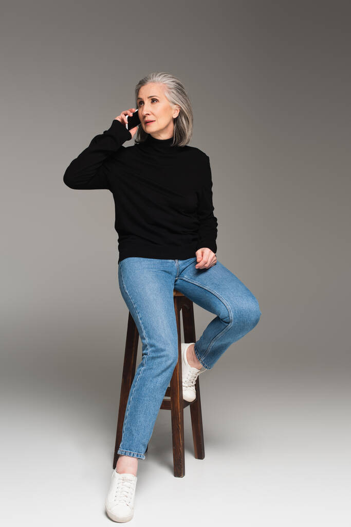 Siwe włosy kobieta mówi na smartfonie na krześle na szarym tle  - Zdjęcie, obraz