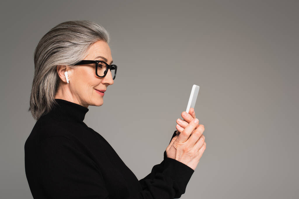 Donna matura in auricolare utilizzando smartphone isolato su grigio  - Foto, immagini