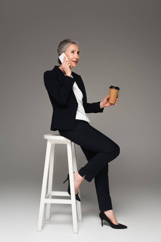 Усміхнена бізнес-леді з кавою, що розмовляє на мобільному телефоні на сірому фоні
  - Фото, зображення