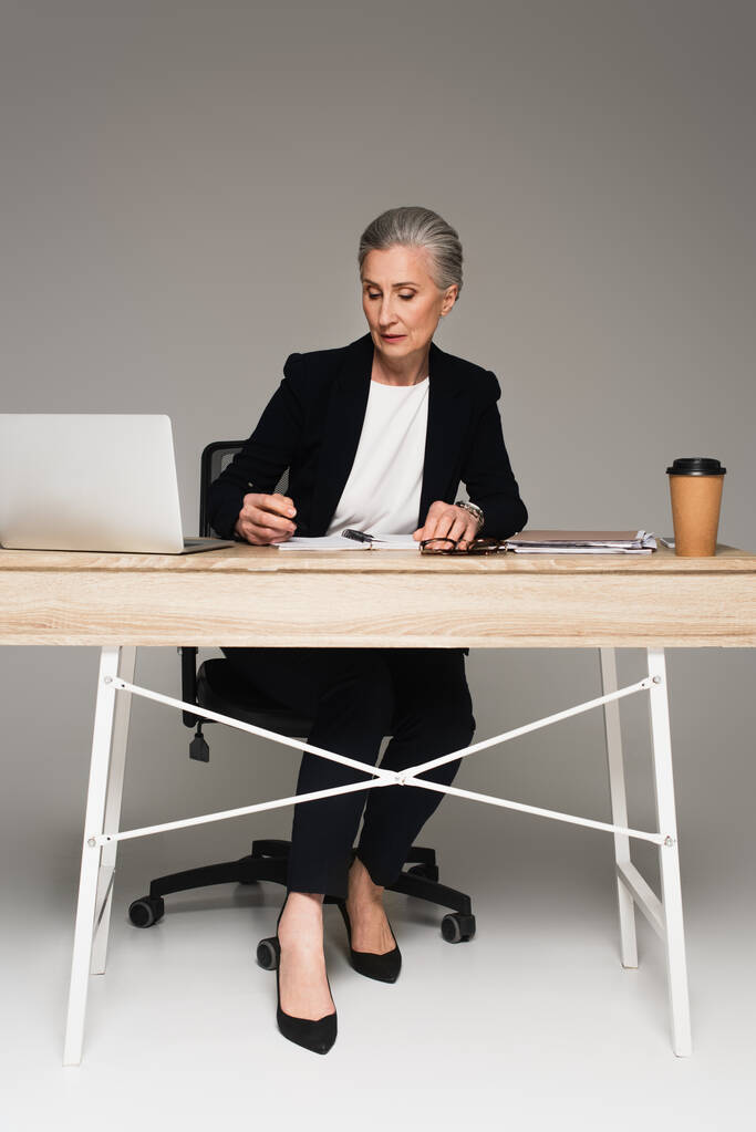 Зрелая деловая женщина смотрит на ноутбук рядом с устройствами и кофе, чтобы пойти на сером фоне  - Фото, изображение