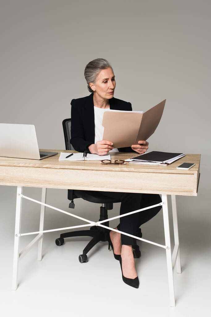 Reife Geschäftsfrau betrachtet Dokumente in der Nähe von Gadgets und Brillen auf einem Tisch auf grauem Hintergrund  - Foto, Bild