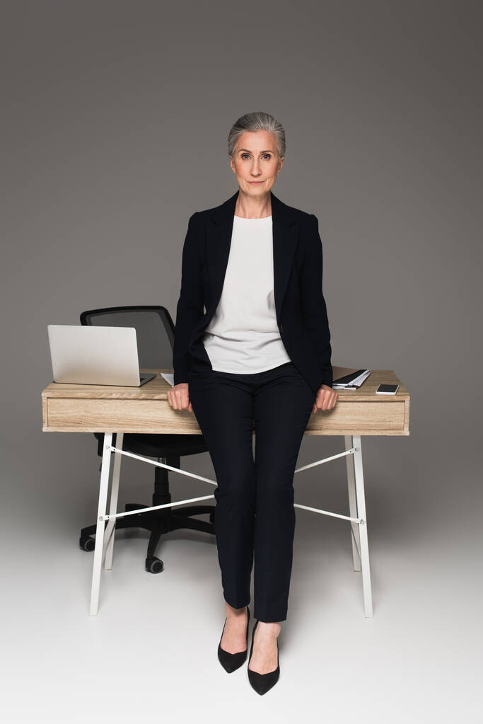 Reife Geschäftsfrau schaut auf Kamera in der Nähe von Gadgets auf Tisch auf grauem Hintergrund  - Foto, Bild