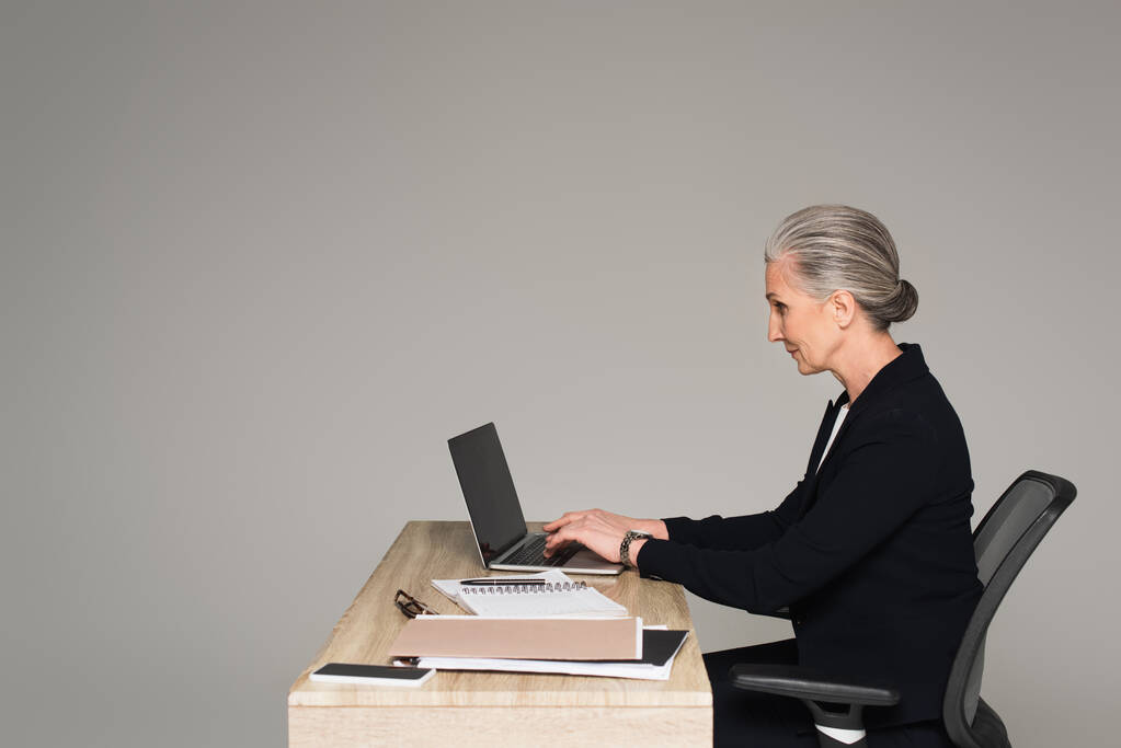 Vista lateral de la mujer de negocios utilizando el ordenador portátil cerca de papeles aislados en gris  - Foto, imagen