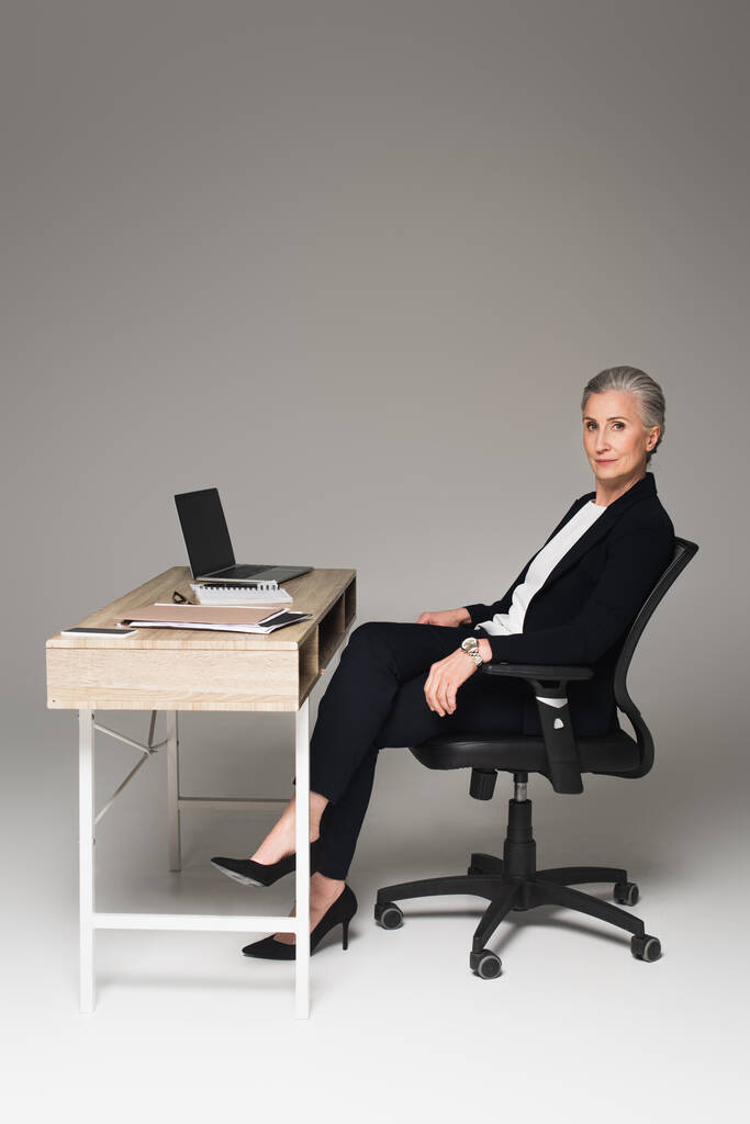 Femme d'affaires mature assise près de gadgets et de papiers sur la table sur fond gris  - Photo, image
