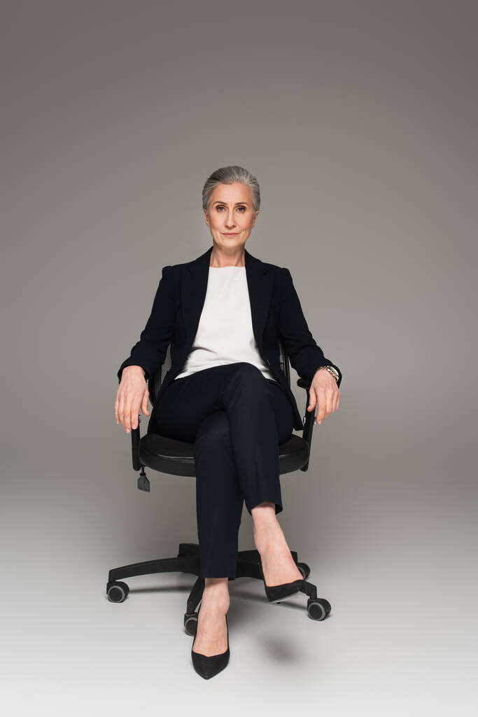 Зрелая деловая женщина смотрит в камеру на офисном стуле на сером фоне  - Фото, изображение