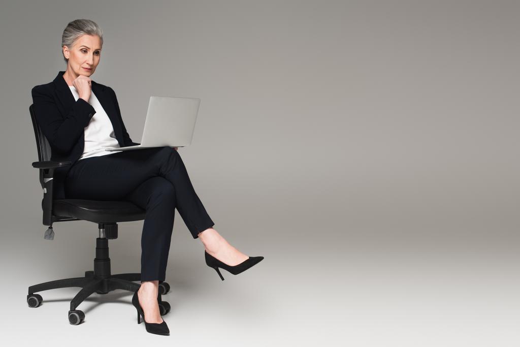 Manažer středního věku při pohledu na notebook na kancelářské židli na šedém pozadí  - Fotografie, Obrázek