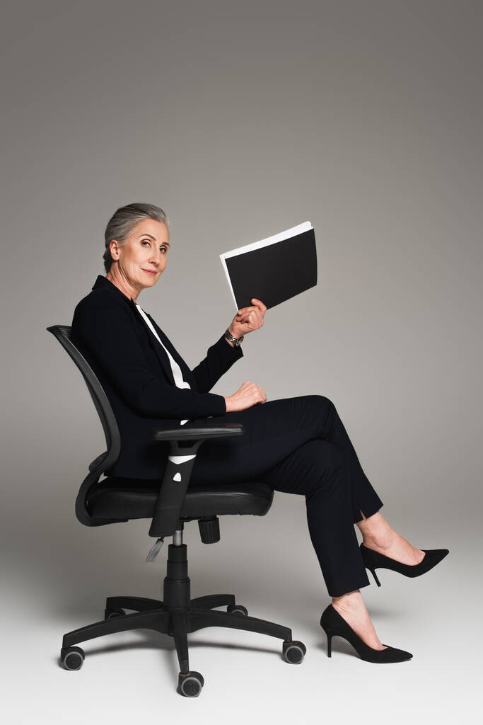 Dojrzała przedsiębiorczyni trzymająca teczkę papierową na krześle biurowym na szarym tle  - Zdjęcie, obraz
