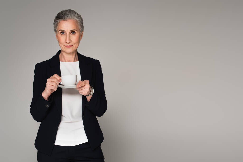 Donna d'affari dai capelli grigi che tiene una tazza di caffè isolata su grigio  - Foto, immagini