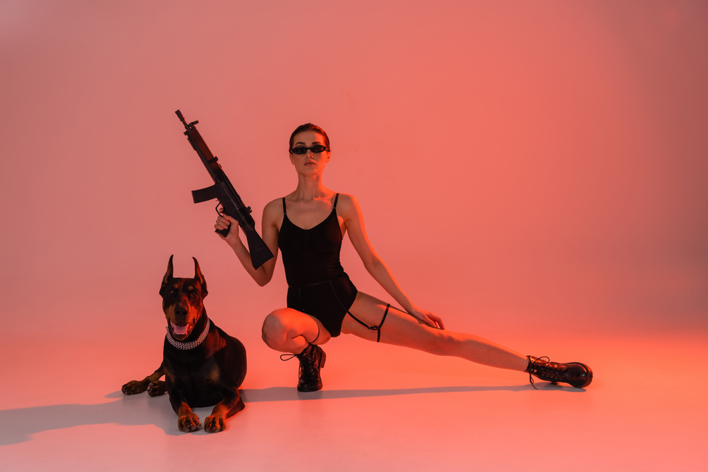 сексуальная женщина позирует с винтовкой рядом с собакой добермана на розовом фоне с желтым светом - Фото, изображение