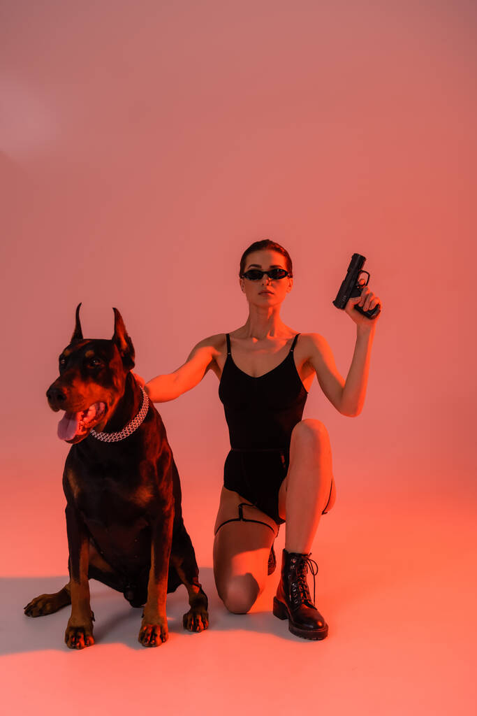 Доберман собака в ланцюговому комірці біля сексуальної жінки з пістолетом на рожевому фоні з жовтим світлом
 - Фото, зображення