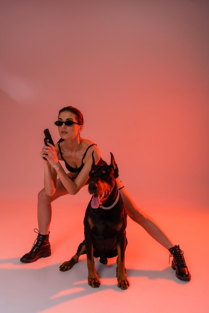 黄色の光とピンクの背景にボディスーツとサングラスを着た武装した女性の近くのドーバーマン犬 - 写真・画像