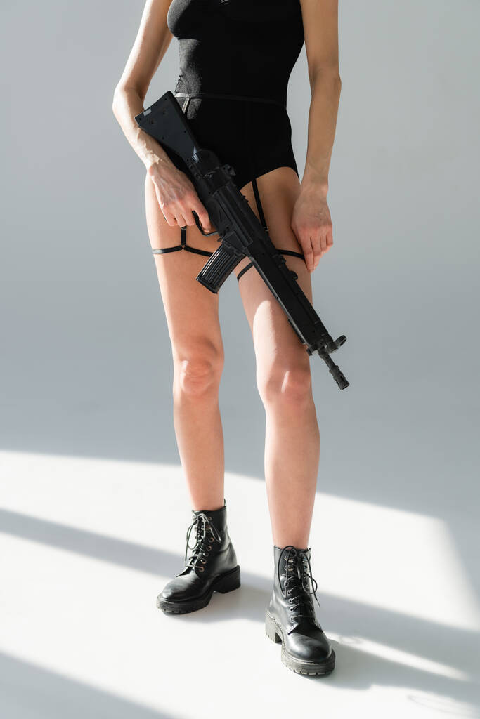 abgeschnittene Ansicht einer Frau in Lederstiefeln mit Gewehr auf grauem Hintergrund mit Schatten - Foto, Bild