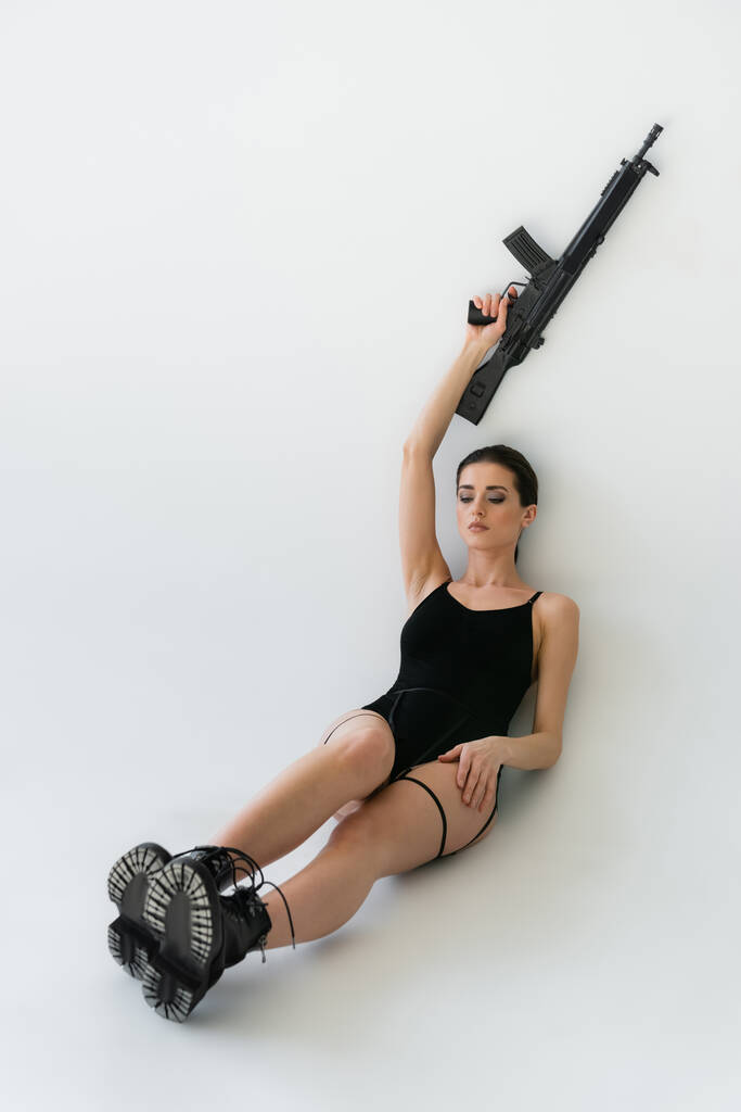 sexy woman in black bodysuit and boots holding rifle in raised hand while sitting on grey  - Photo, Image