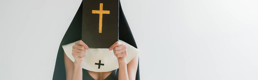 vista recortada de monja sexy que oscurece la cara con la Biblia aislado en gris, bandera - Foto, Imagen