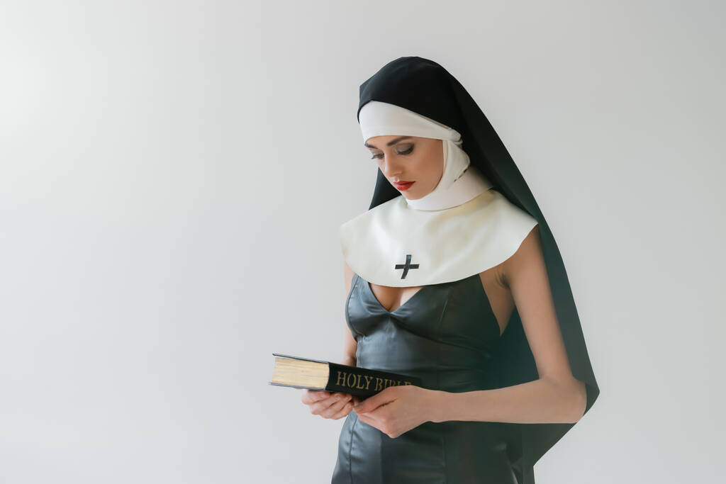 νεαρή καλόγρια με σέξι φόρεμα που κρατά την Αγία Γραφή απομονωμένη στο γκρι - Φωτογραφία, εικόνα