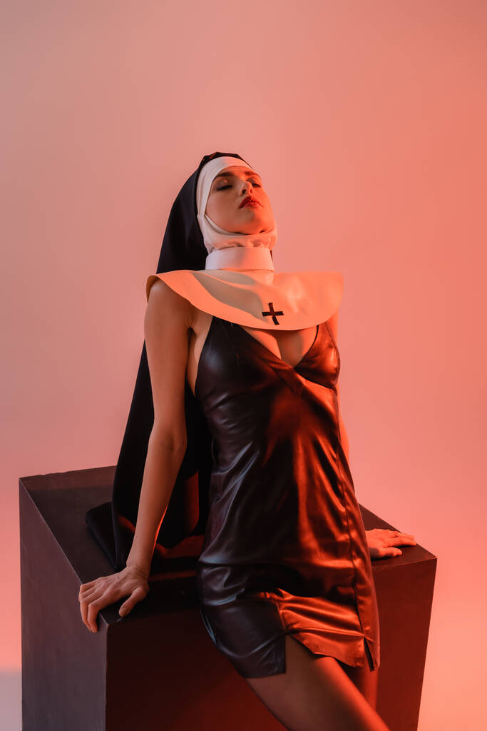 érzéki apáca szexi ruha pózol csukott szemmel közelében fekete kocka rózsaszín háttér - Fotó, kép
