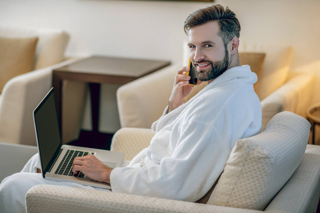 Homem barbudo jovem em um manto de banho branco sentado com um laptop - Foto, Imagem