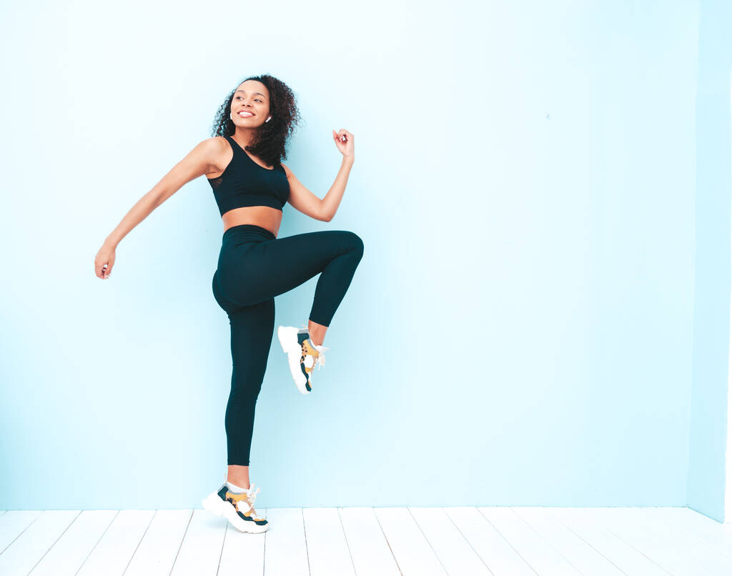 Fitness mosolygó fekete nő sport ruházat afro fürtök frizura.Ő visel sportruházat. Fiatal gyönyörű modell tökéletes cserzett test.Nő ugrás és fut a stúdióban, közel a kék fal - Fotó, kép