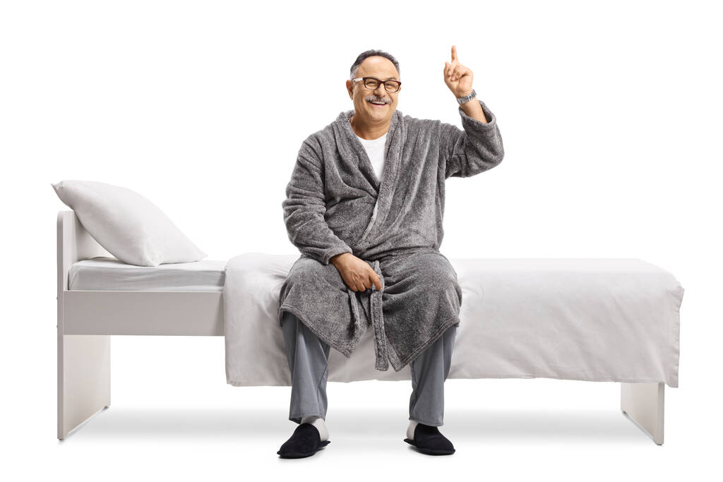 Homme mûr souriant en pyjama et une robe assise sur un lit et pointant vers le haut isolé sur fond blanc - Photo, image