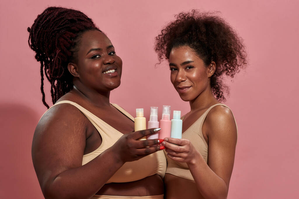 Zwei junge Afro-Frauen tragen beige Unterwäsche und halten Flaschen mit Pflegeprodukten in der Hand - Foto, Bild