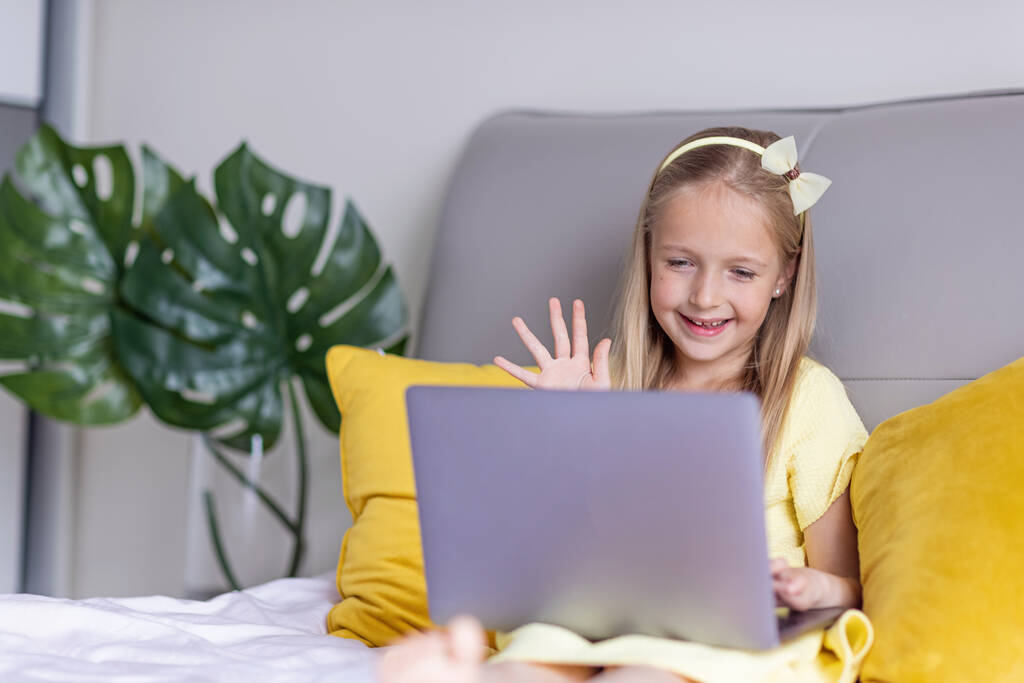 Χαριτωμένο μικρό καυκάσιο κορίτσι με ξανθά μαλλιά σε μοντέρνο φόρεμα που φωτίζει το κίτρινο χρώμα κάθεται στο σπίτι κατά τη διάρκεια της πανδημίας coronavirus καραντίνα και τη χρήση φορητού υπολογιστή. Μείνετε στο σπίτι κατά τη διάρκεια covid-19 - Φωτογραφία, εικόνα
