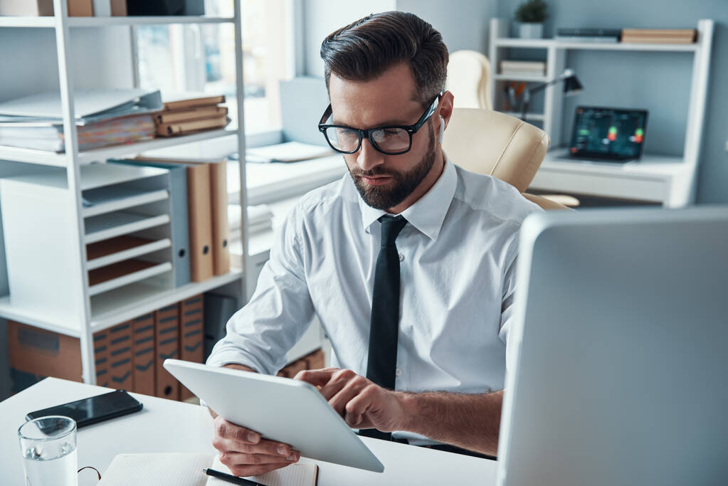 Zajęty młody człowiek w koszuli i krawacie pracujący przy użyciu technologii bezprzewodowych siedząc w biurze - Zdjęcie, obraz