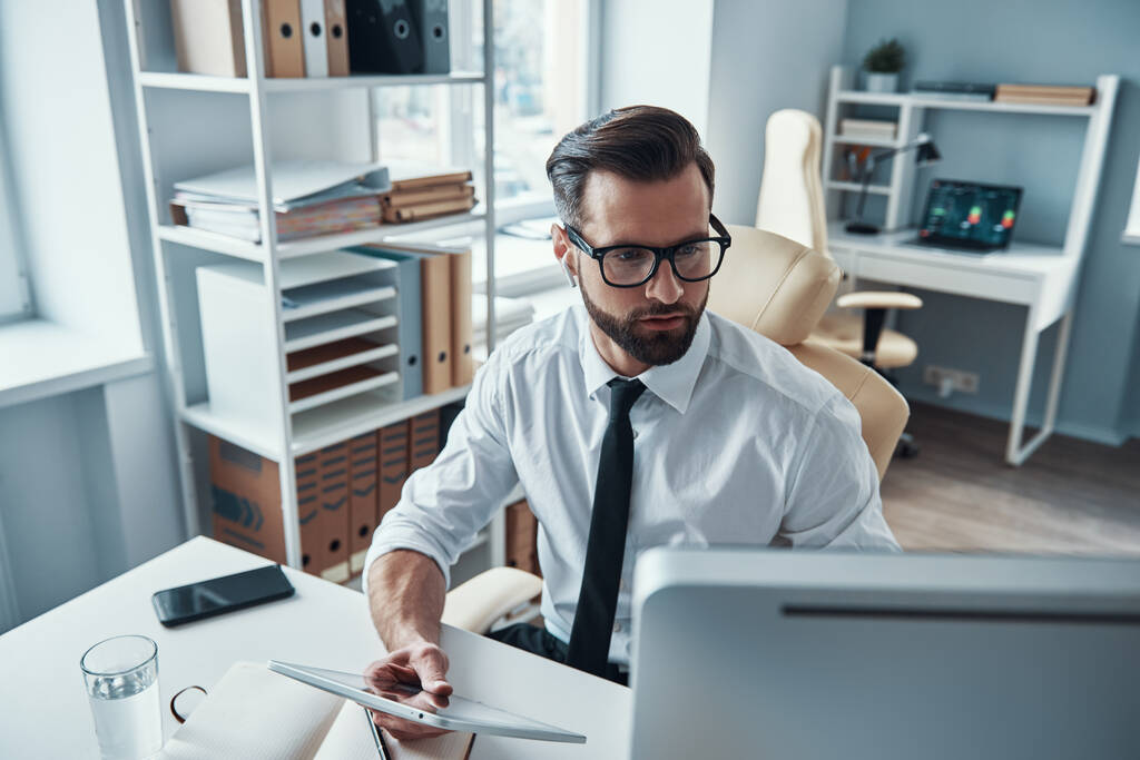 Gömlek ve kravatlı konsantre genç adam ofiste otururken kablosuz teknoloji kullanarak çalışıyor. - Fotoğraf, Görsel