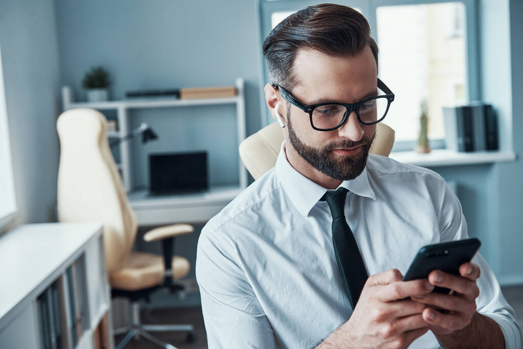 Красивый молодой человек в формальной одежде с помощью смартфона и наушников, сидя в офисе - Фото, изображение