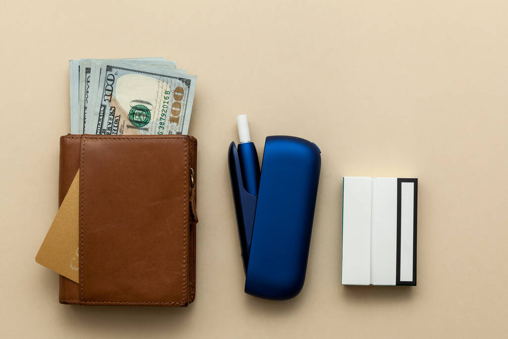 Elektroniczny niebieski papieros IQOS, brązowy portfel z pieniędzmi na beżowym tle. Zdjęcie z miejscem na tekst, logo i projekt. Odpoczynek koncepcyjny. - Zdjęcie, obraz