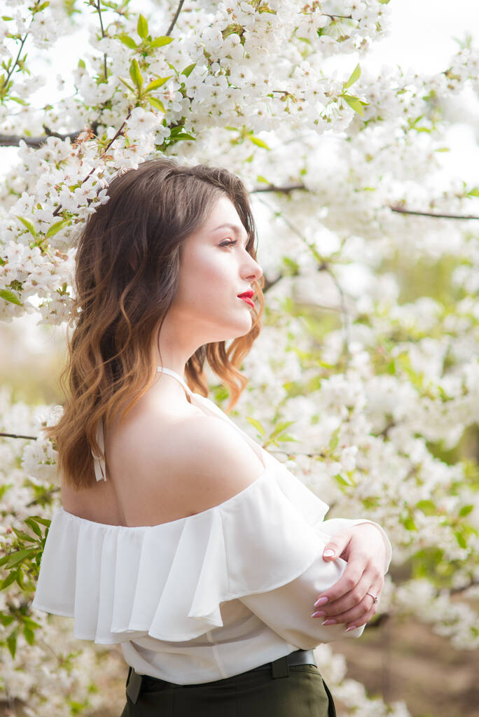 Ihana nuori tyttö valkoisessa romanttisessa silkkipuserossa keväällä kukkivassa kirsikkapuutarhassa aurinkoisella säällä. Kevät ja muoti
 - Valokuva, kuva