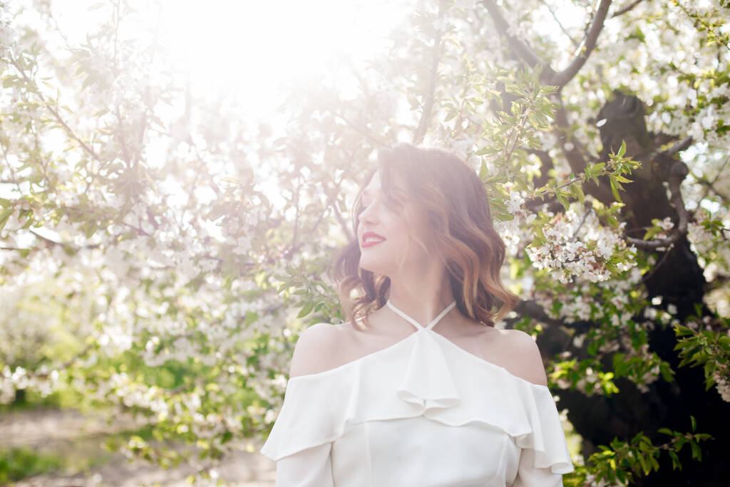 Menina encantadora em uma blusa de seda romântica branca na primavera em um jardim de cereja florescente em tempo ensolarado. Primavera e moda
 - Foto, Imagem