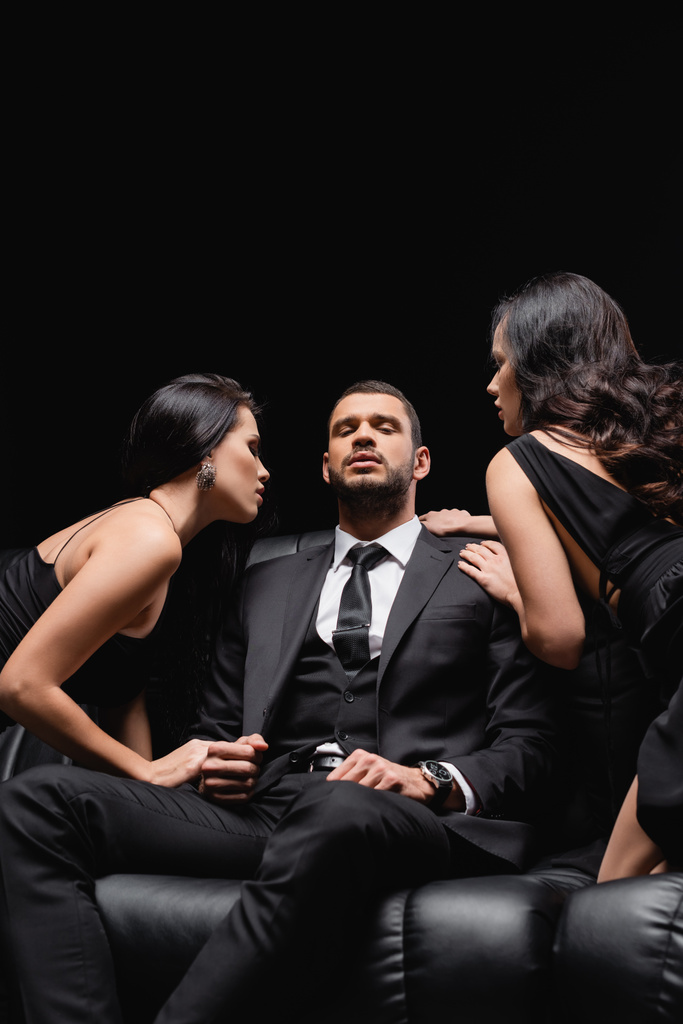 uomo d'affari di successo seduto sul divano di pelle vicino alle donne seducendolo isolato su nero - Foto, immagini