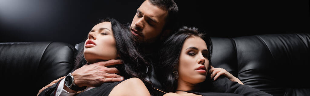joven abrazando a mujeres seductoras mientras está sentado en el sofá de cuero en negro, pancarta - Foto, imagen