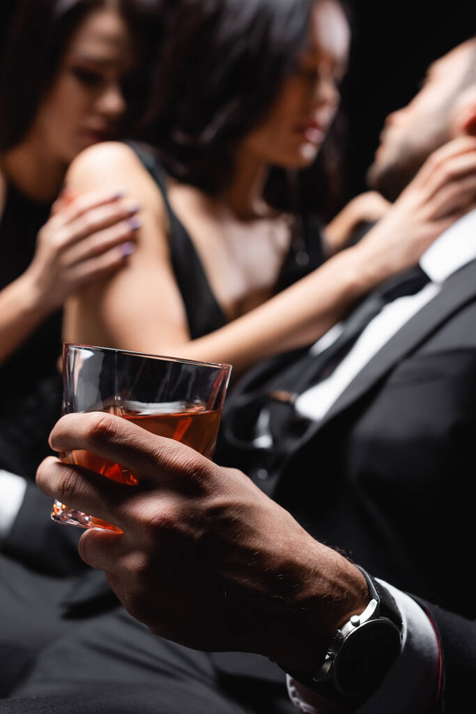 Tutkulu kadınların yanında viski bardağı taşıyan bir adam bulanık arka planda, siyahta izole edilmiş. - Fotoğraf, Görsel