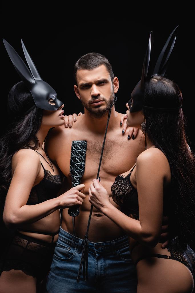 passionate women in bunny masks holding flogging whip and spanking paddle near sexy man isolated on black - Photo, Image