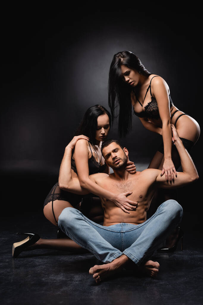 сексуальный мужчина сидит со скрещенными ногами и смотрит в камеру рядом с чувственными женщинами на черном фоне - Фото, изображение