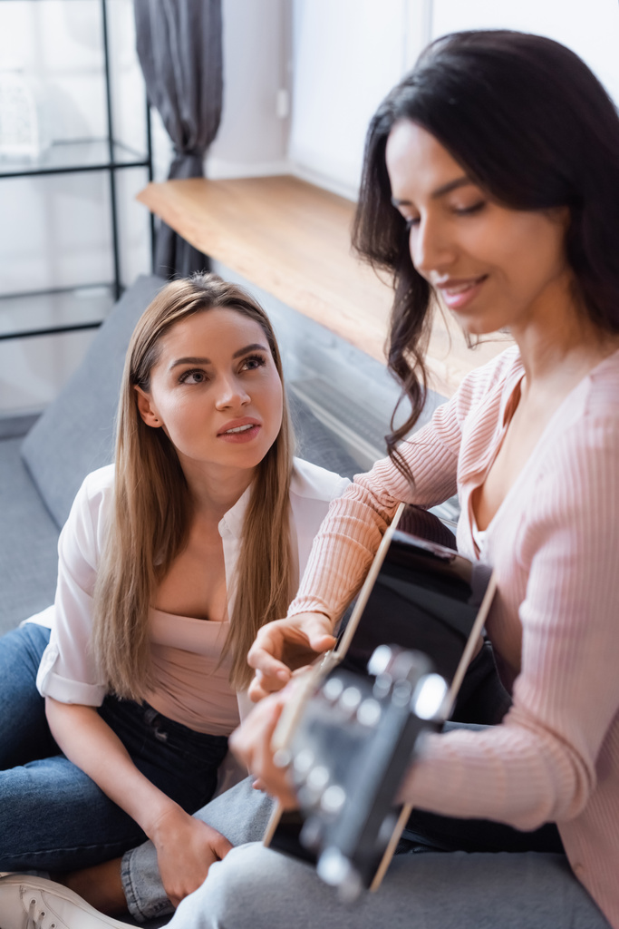 homályos barna nő játszik akusztikus gitár közelében barátnője a nappaliban  - Fotó, kép