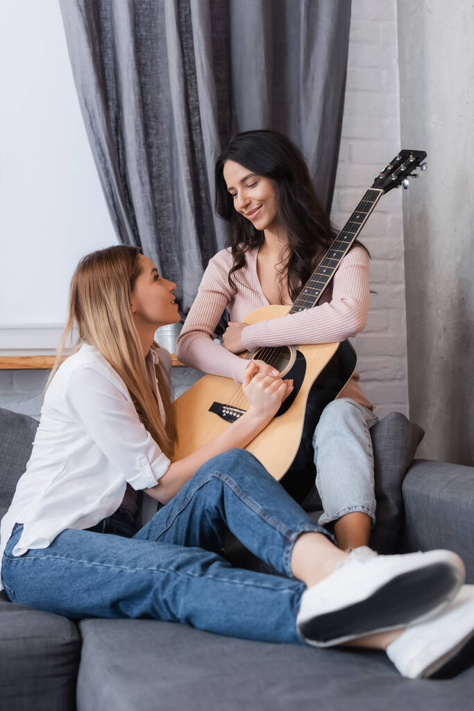 brunette vrouw en vrolijke vriendin houden handen in de buurt van akoestische gitaar in de woonkamer  - Foto, afbeelding