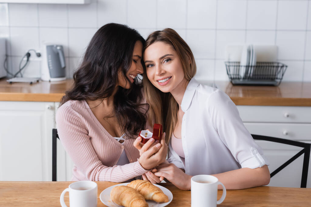 smiling woman holding jewelry box with wedding ring near girlfriend and breakfast in kitchen  - Foto, Imagen
